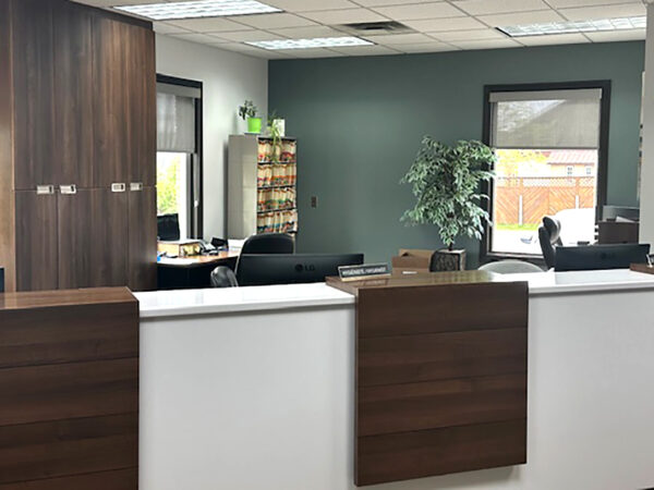 A modern dental office reception area featuring a wooden and white front desk, comfortable chairs, and plants. The space is well-lit with large windows and has a welcoming atmosphere. The 123Dentist Smiley is present, adding a friendly touch to the environment.