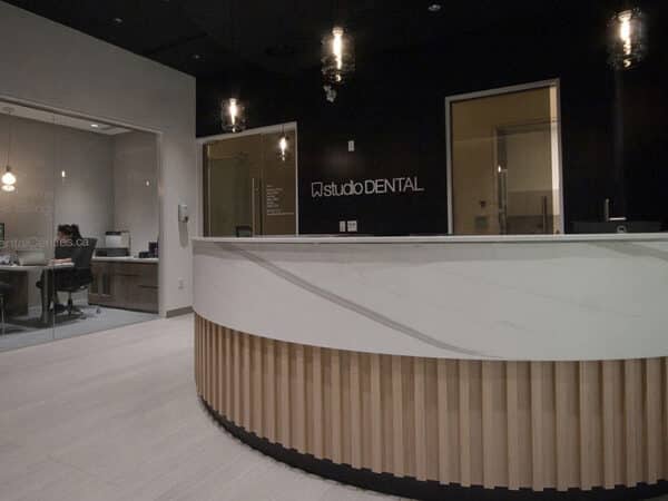 A modern dental office reception area featuring a curved wooden front desk, warm lighting, and a dark wall with the logo "Cloverdale DENTAL." In the background, dental staff can be seen working at their stations, creating a welcoming atmosphere.