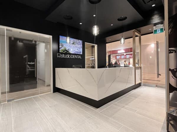 A modern dental clinic reception area featuring a sleek, minimalist design with a light-colored front desk, dark walls, and a digital display. The setting is well-lit and inviting, perfect for welcoming patients. The friendly 123Dentist Smiley adds a cheerful touch to the atmosphere.