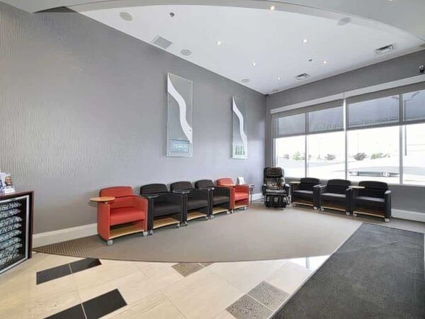 A modern waiting area features a gray textured wall, two abstract artworks, red and black seating, and large windows allowing natural light. The floor has a stylish pattern, creating a welcoming atmosphere.