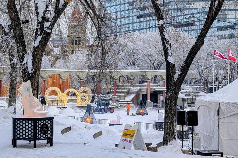 A snowy park scene featuring frosted trees, ice sculptures, and colorful decorations. In the background, a building glimmers through the snow, while people engage in winter activities. Flags and tents add to the festive atmosphere.