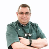 A smiling dentist in a green coat stands with arms crossed, wearing glasses and dental tools around his neck. The background is plain white, emphasizing his friendly demeanor.