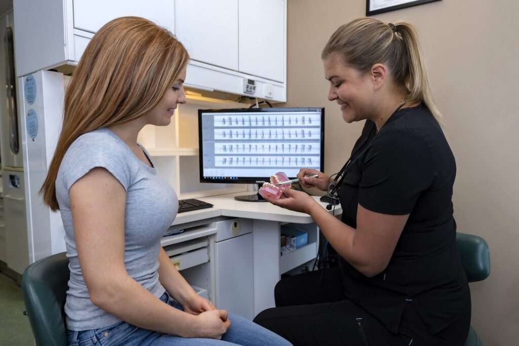 A dental professional discusses oral health with a patient in a clinic setting. The hygienist holds a dental model, while the patient listens attentively. A computer screen displaying dental information is visible in the background. The friendly 123Dentist Smiley adds a cheerful vibe to the environment.