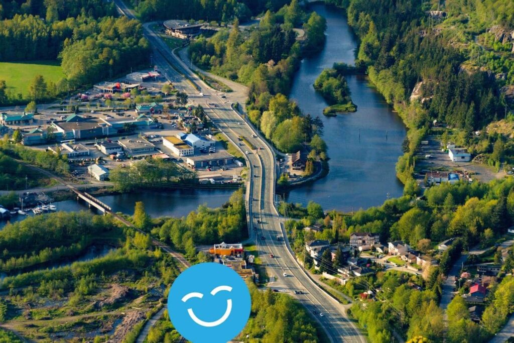 Aerial view of a lush landscape featuring winding roads, a river, and clusters of green trees. Nearby, residential and commercial buildings are visible, surrounded by nature. A cheerful graphic is superimposed at the bottom.