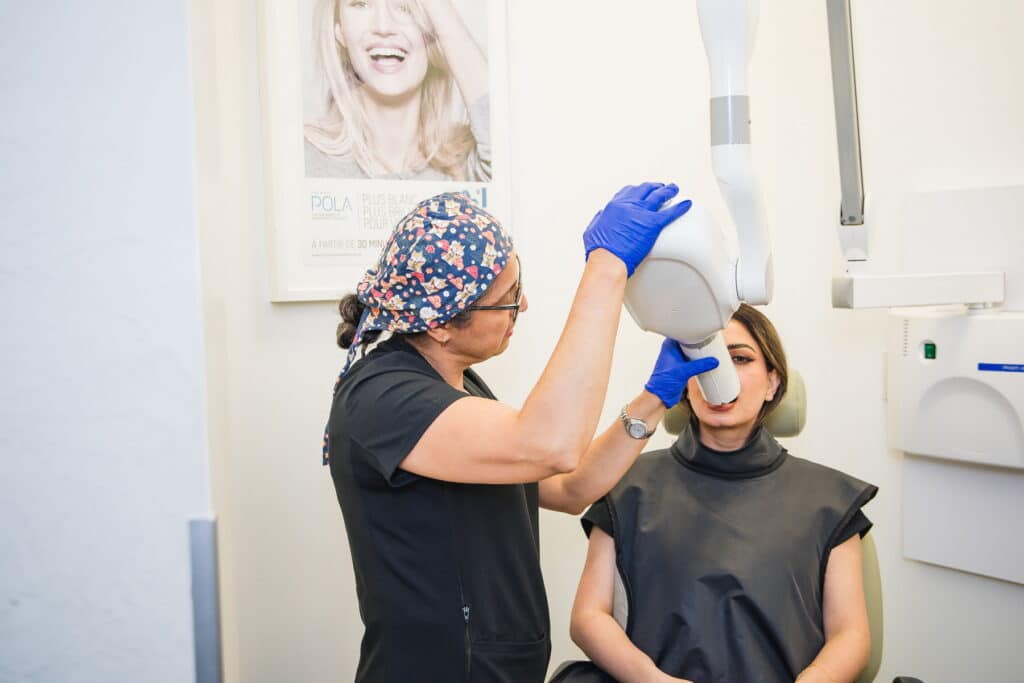 A dental professional wearing gloves adjusts equipment while a patient sits in a dental chair, prepared for a procedure. A cheerful poster of a smiling woman is visible in the background.