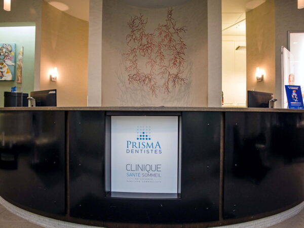 A modern reception desk at Prisma Clinics, featuring a central logo and soft lighting. The wall behind has an artistic design, creating a welcoming atmosphere for visitors.