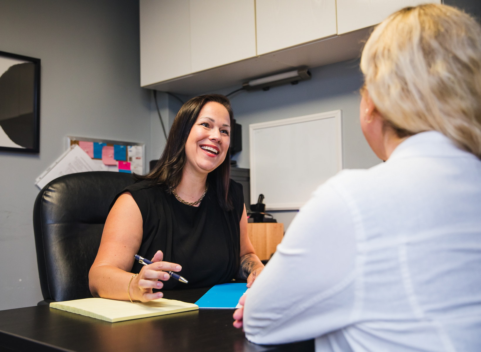 A woman in a black blouse sits at a desk, smiling and engaging in conversation with another woman. The setting appears to be an office, with documents and notes visible on the desk.