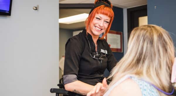 A cheerful dental professional with bright orange hair smiles while interacting with a patient in a dental office setting. The atmosphere is friendly and welcoming, making the patient feel at ease. The 123Dentist Smiley can be seen in the background, adding to the positive vibe.