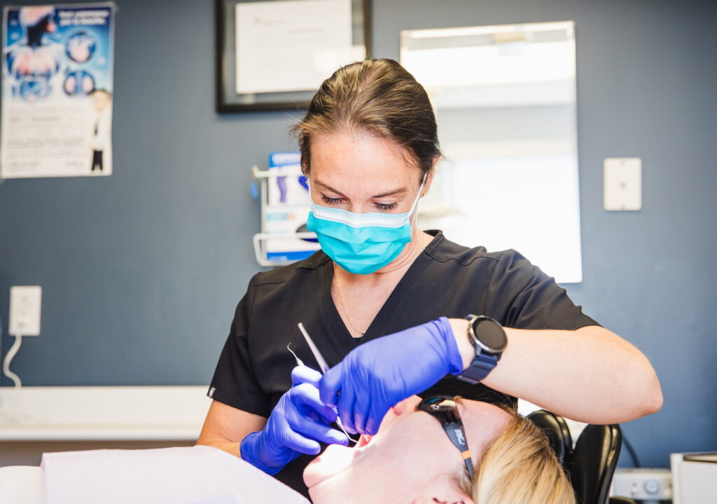 A dental hygienist in scrubs and a mask is providing an oral examination to a patient in a dental office. The patient is reclining in a chair and wearing protective glasses while the hygienist uses dental tools.