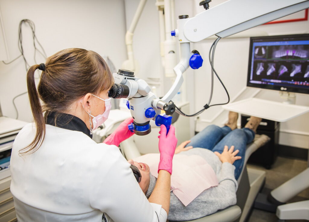 A dental professional in gloves uses a microscope to examine a patient lying down in a dental office. Medical equipment and a monitor displaying images of teeth are visible in the background, creating a clinical environment focused on dental care. 123Dentist Smiley adds a cheerful touch to the scene.