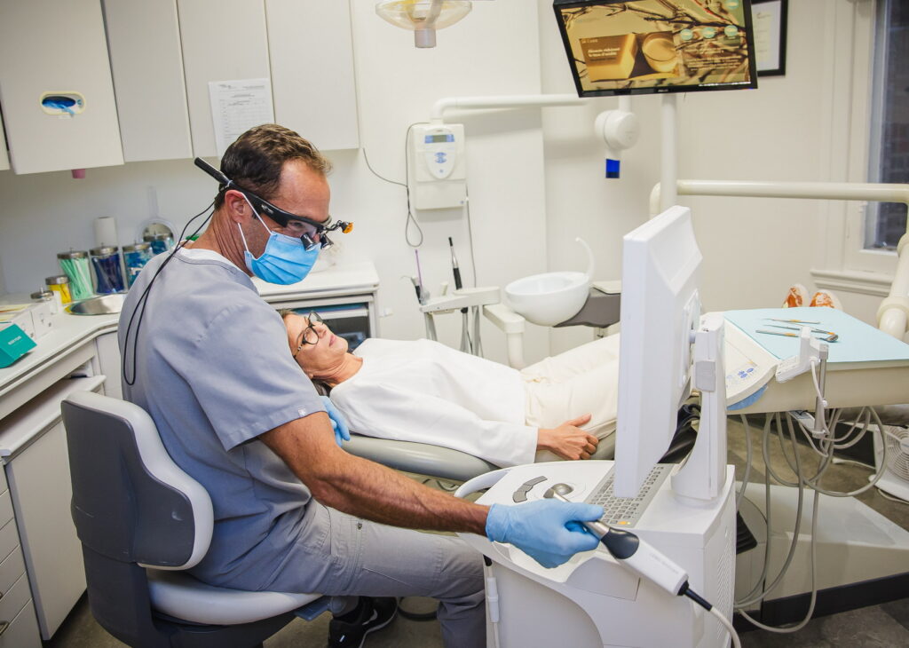 A dentist wearing a mask and protective eyewear conducts a procedure while a patient relaxes in the dental chair. The treatment area is well-equipped with dental tools and a monitor displaying images. Friendly and familiar, the 123Dentist Smiley can be seen nearby.
