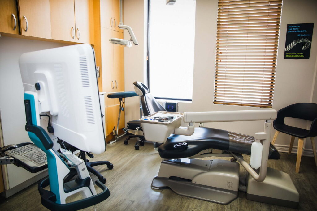 A dental clinic interior featuring a dental chair, computer workstation, and tools. The room has wooden cabinets, a chair, and natural light from a window with blinds, creating a clean and professional atmosphere.