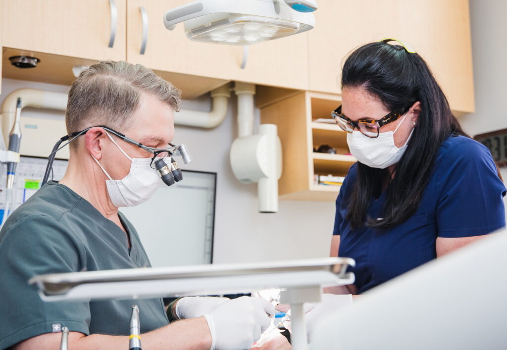 A dental professional wearing magnifying glasses collaborates with an assistant in a clinic. Both are wearing masks, focusing on a patient care procedure. The atmosphere reflects a clean, efficient dental environment.