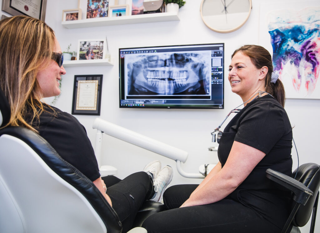 A dental professional speaks with a patient in a modern dental office. An X-ray is displayed on a monitor behind them, highlighting the patient's dental health. Both individuals are wearing black scrubs and appear engaged in a friendly conversation. The 123Dentist Smiley adds a cheerful touch to the environment.