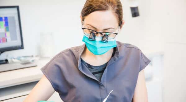 A dental professional wearing a mask and protective glasses diligently examines a patient's teeth in a bright clinic setting. Tools are in hand, and equipment is visible in the background, highlighting a focus on dental care and hygiene.