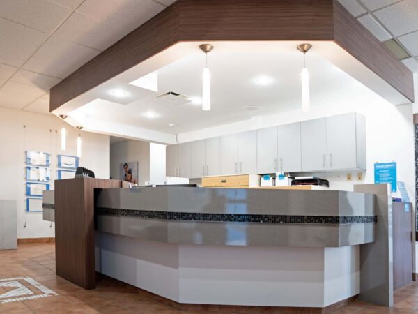 A modern reception area featuring a curved front desk with a sleek design, light wood accents, and pendant lighting. The background shows a bright space with gray cabinetry and informational displays, creating an inviting atmosphere for visitors.