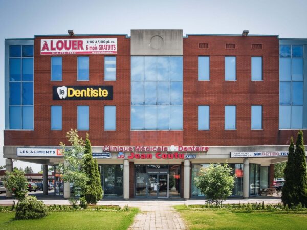 A modern brick building featuring multiple storefronts, including a dental office, set in a landscaped area with grass and small trees. Large signs display business names and rental information, indicating the building's commercial purpose.