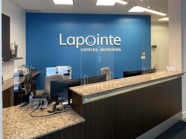 A modern dental office reception area featuring a granite counter, sleek chairs, and a blue wall with the "Lapointe centres dentaires" sign. The space is bright and welcoming, designed for patient comfort and ease of access.