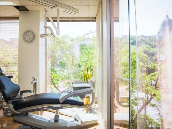 A bright dental office featuring a comfortable examination chair, large windows with views of greenery, and a wall clock. Natural light illuminates the space, creating a welcoming atmosphere.