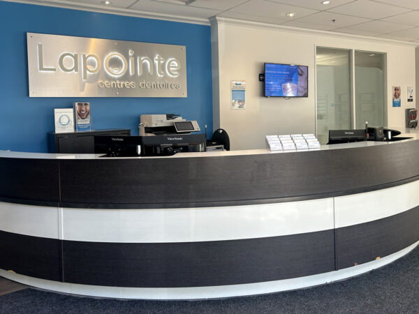 A modern dental office reception area featuring a curved front desk with a black and white design. The wall behind is painted blue with the sign "Lapointe.” A television is mounted on the wall, and informational materials are available on the desk. The friendly 123Dentist Smiley adds a welcoming touch.