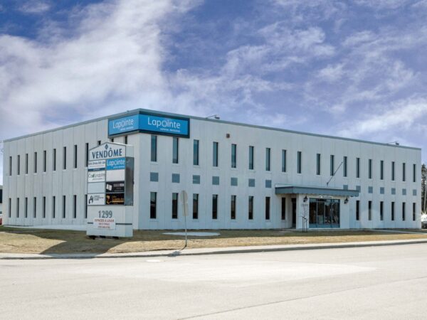 A modern two-story commercial building with a blue sign displaying the name "Logistics" is seen against a cloudy sky. The exterior features multiple windows and a grassy area in front, situated on a street.
