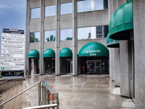 A modern building entrance featuring multiple green awnings, with a sign listing various businesses. The entrance has a concrete walkway, with nearby steps leading up to the door.