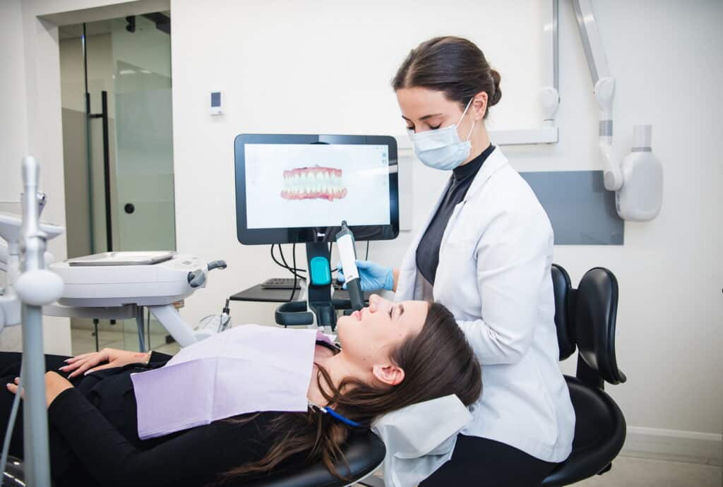 A dental professional in a white coat assists a patient seated in a chair. The patient is reclined with a dental bib, while a digital screen displaying dental images is visible in the background. The setting suggests a modern dental clinic focused on oral care.