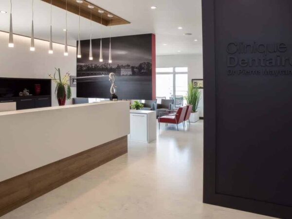 A modern dental clinic reception area featuring a white reception desk, stylish seating, decorative plants, and contemporary lighting. The background includes a large wall photo and a sign indicating the clinic's name. The space is bright and inviting, designed for patient comfort.