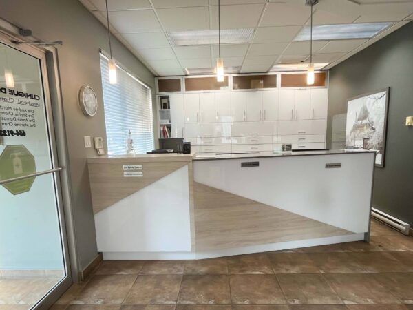 A modern reception area featuring a minimalist front desk with a geometric design, light-colored cabinets, and large windows allowing natural light. The walls are painted a soft gray, creating a welcoming atmosphere.