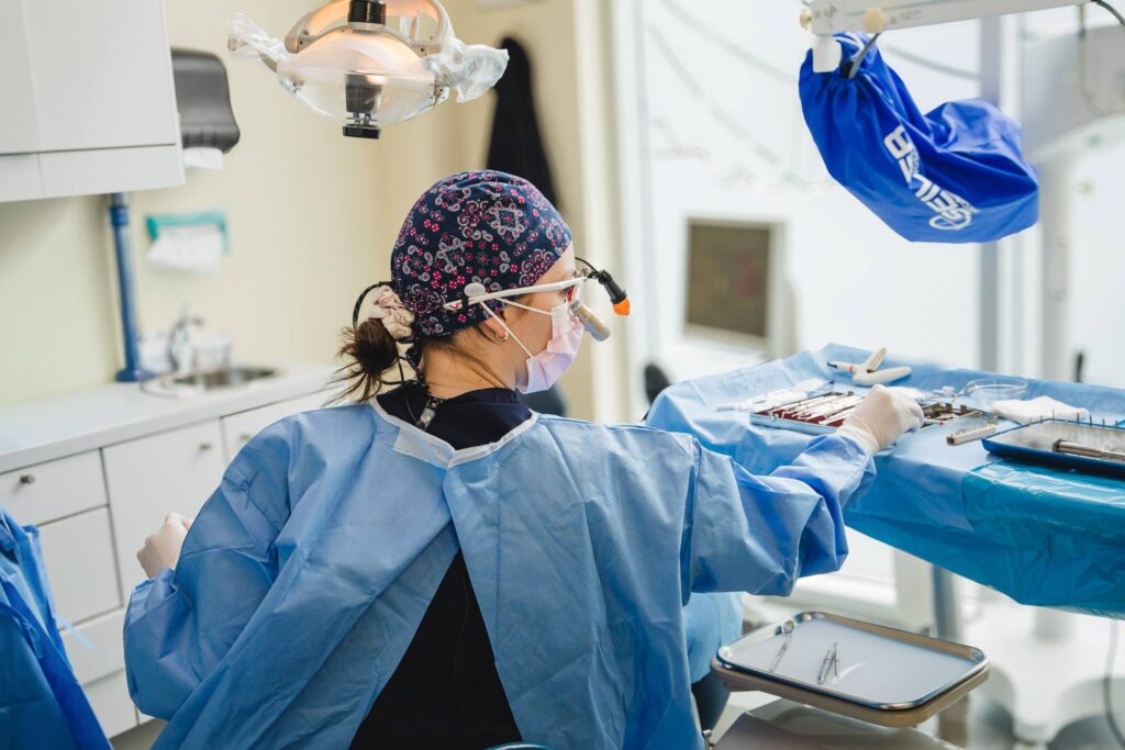 A dental professional, wearing scrubs and a surgical mask, is focused on a procedure in a well-lit clinic. Instruments are laid out on a blue drape, and dental equipment is visible in the background. The 123Dentist Smiley adds a friendly touch to the dental environment.