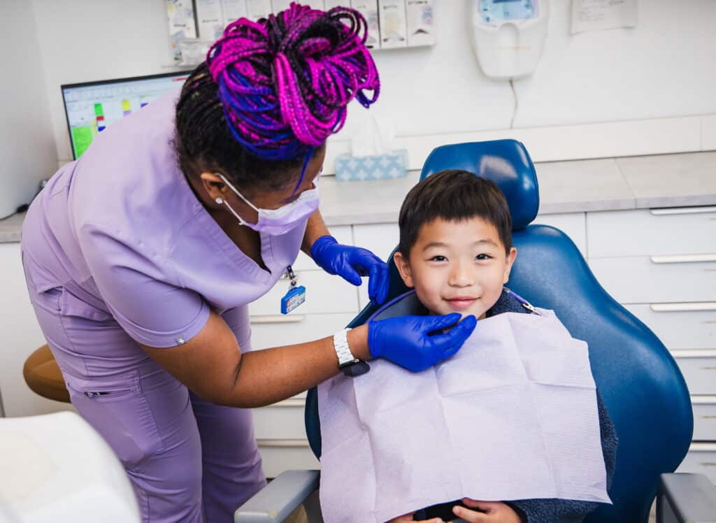 A dental professional in purple attire is gently preparing a young boy for a procedure in a dental office. The boy smiles while seated in a dental chair, wearing a protective bib. A friendly blue 123Dentist Smiley is also present, adding a cheerful vibe to the scene.