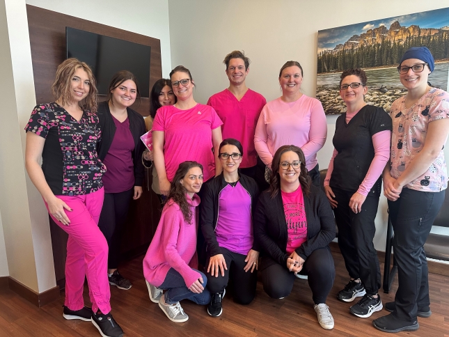 A team of dental professionals poses together in a brightly lit office, wearing various shades of pink. They display a friendly atmosphere and camaraderie, ready to assist patients. In the background, there is a scenic landscape painting.