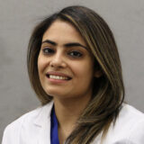 A smiling woman with long, wavy hair wearing a white lab coat poses in front of a neutral background. She exudes a friendly and professional demeanor, likely representing a healthcare or dental setting.