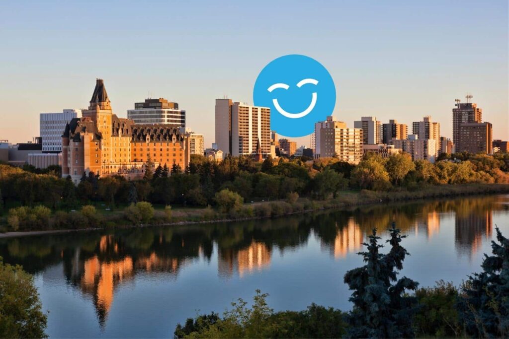 A scenic view of a city skyline at sunset, featuring modern buildings and a historic structure reflected in the calm river. Lush trees frame the foreground, enhancing the tranquil atmosphere. A cheerful blue smiley icon is superimposed on the image.