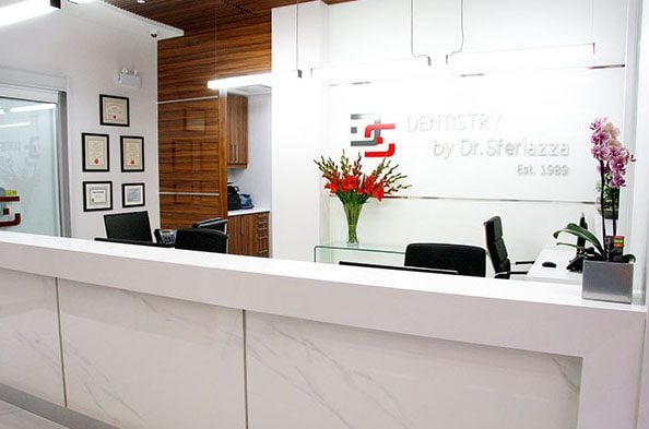 Modern dental office reception area featuring a sleek white desk, certificates on the wall, and a vase of flowers. The design is clean and inviting, with warm wooden accents and contemporary lighting.