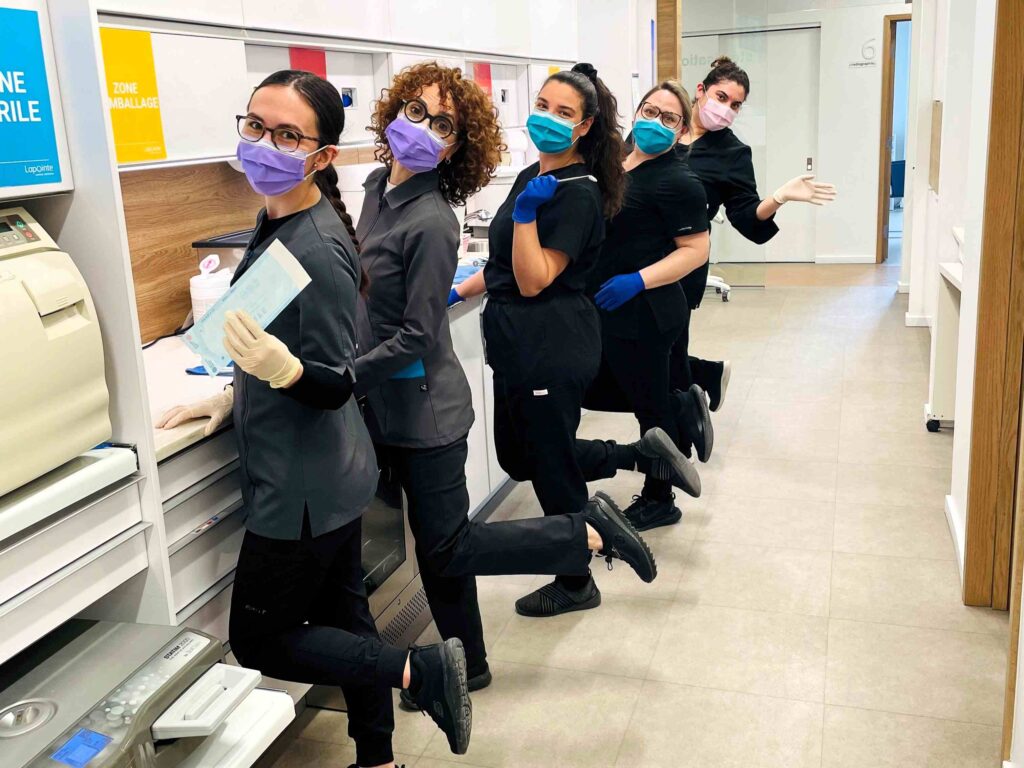 A group of five dental professionals wearing masks and gloves pose playfully in a bright, modern dental office. They stand in a line, smiling and showcasing their teamwork. The friendly 123Dentist Smiley adds a cheerful vibe to the scene.