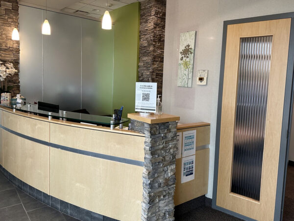 A modern dental clinic reception area featuring a curved wooden front desk with a stone accent. Soft lighting and green décor create a welcoming atmosphere. Nearby, a glass door leads to treatment areas, and informational displays are present. The friendly 123Dentist Smiley adds a cheerful touch to the space.