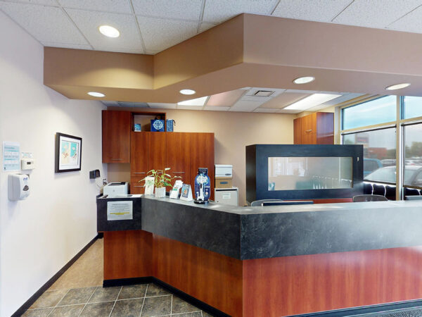 A bright and welcoming dental office reception area featuring a sleek front desk, wooden cabinets, and large windows. The space is organized with dental materials and decor, creating a friendly environment for patients. The 123Dentist Smiley adds a cheerful touch to the atmosphere.