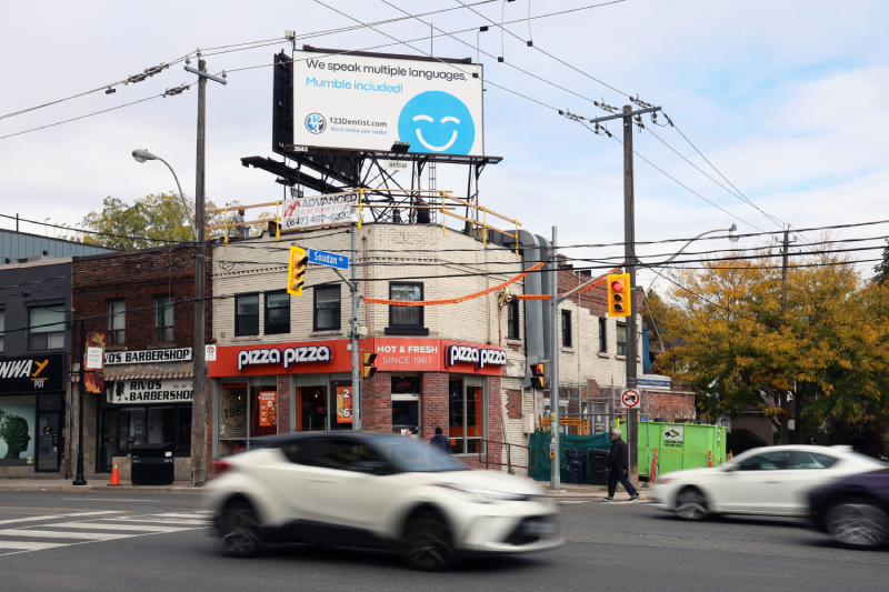 A street scene features a pizza restaurant at the corner with a billboard advertising dental services, featuring the friendly 123Dentist Smiley. Cars are passing by, and trees with autumn leaves provide a colorful backdrop.