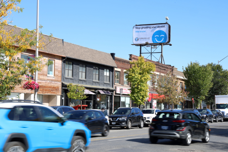 A busy street scene features cars passing by quaint shops and vibrant trees. Prominently displayed is a billboard featuring the friendly 123Dentist Smiley against a clear blue sky, inviting community engagement.