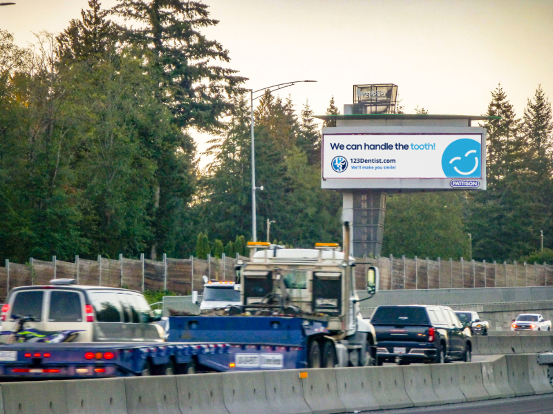 A busy highway scene featuring various vehicles, with a large billboard displaying the message "We can handle the smiles" alongside the friendly 123Dentist Smiley logo. Surrounding trees indicate a natural setting.