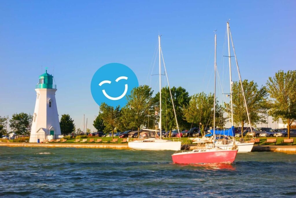 A scenic view of a lighthouse beside calm waters, with a red sailboat anchored nearby. Lush green trees line the shore under a clear blue sky, featuring a smiling icon in the background.