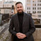 A man in a stylish gray checked blazer and black turtleneck stands outdoors, smiling confidently with his hands clasped. The city skyline is visible in the background, bathed in warm sunset light.