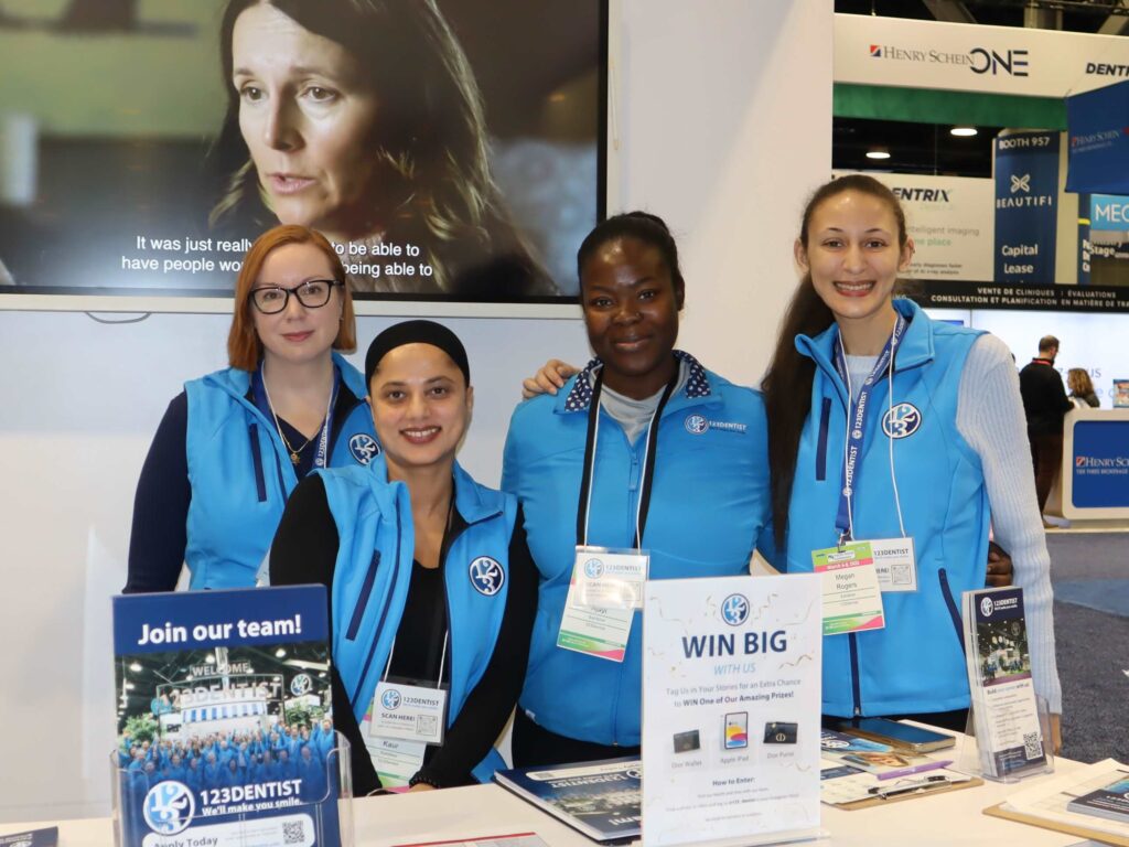 Four individuals in matching blue vests stand at a booth promoting 123Dentist. They smile warmly while showcasing a sign that invites people to join their team. In the background, a soft-focus image of a woman appears, adding a professional atmosphere to the setting.