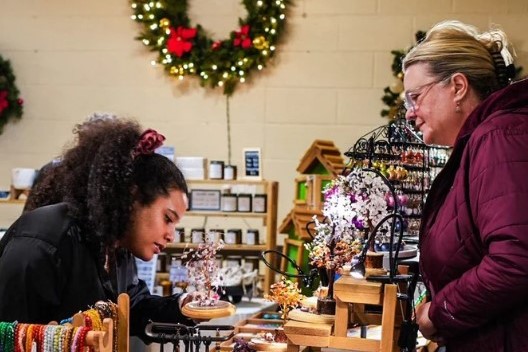 Two women are engaged in a conversation at a retail store, with one woman showing interest in various handmade items. Holiday decorations, including a wreath, are visible in the background, enhancing the festive atmosphere.