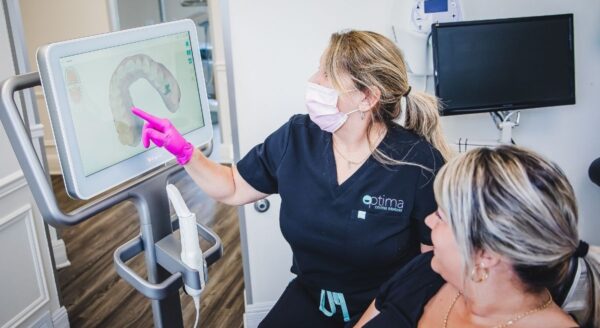 A dental professional shows a patient a digital screen displaying dental information. Both are wearing masks, and the clinician is using pink gloves. The setting is a modern dental office. The friendly 123Dentist Smiley adds a cheerful touch to the environment.