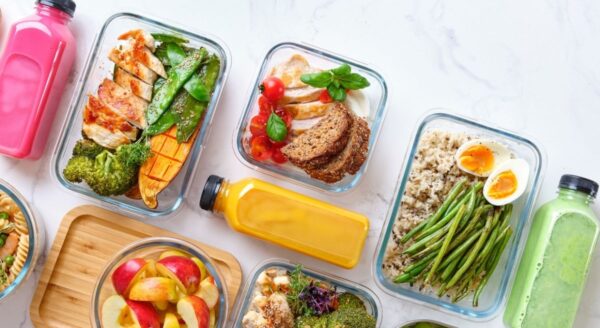 Une collection de contenants colorés pour la préparation de repas remplis d'aliments sains, notamment du poulet grillé, des légumes, des céréales et des fruits. Des bouteilles de smoothies colorés sont disposées à côté, créant une présentation attrayante et nutritive.