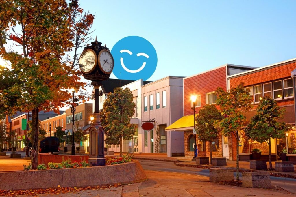 A quaint town square at sunset with a vintage clock, trees, and lit streetlamps lining the street. A smiling blue emoji is overlaid in the sky.