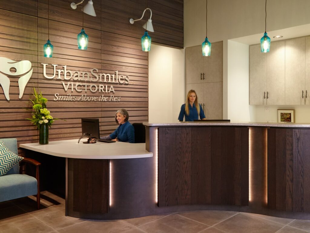 A modern dental reception area at UrbanSmiles Victoria featuring a stylish front desk, two staff members, and decorative lighting. The space is warm and welcoming, designed for patient comfort and care.