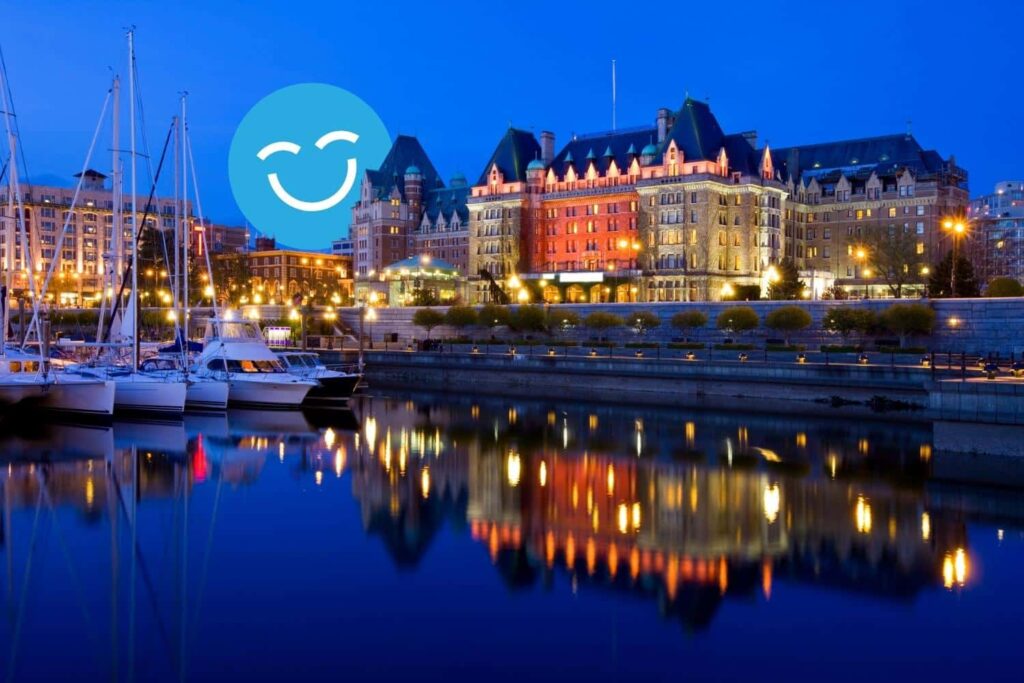 Waterfront view of a lit-up, grand building at dusk, reflecting in the calm water, with docked boats on the left and a smiling blue emoji in the sky.